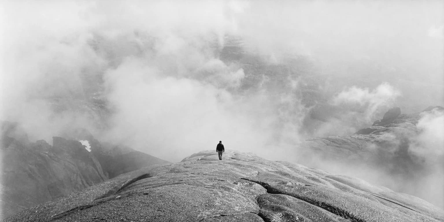 Solitary figure walks toward swirling mountain clouds on a vast granite ridge, black-and-white, evoking quiet resolve and the weight of an irreversible step for