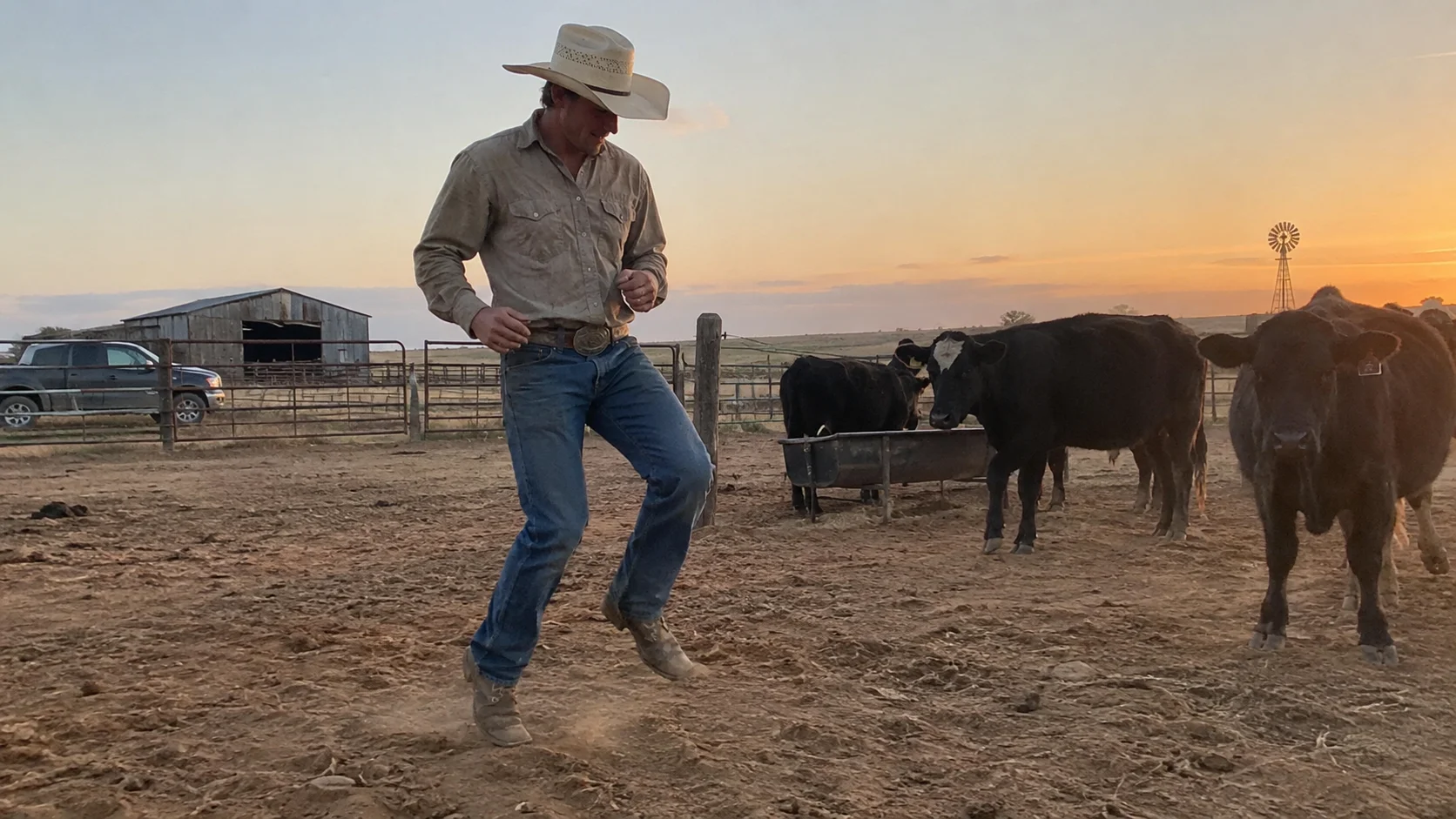 Cowboy in jeans and straw hat strides across a dusty ranch pen at golden-hour sunset, black cattle and a windmill silhouetted behind him.